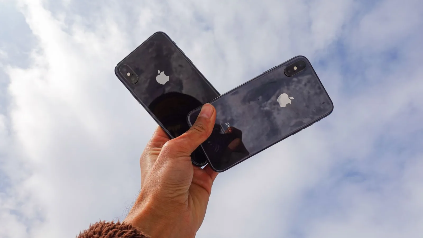Close-up of a hand holding two smartphones against a cloudy sky, showcasing modern technology.