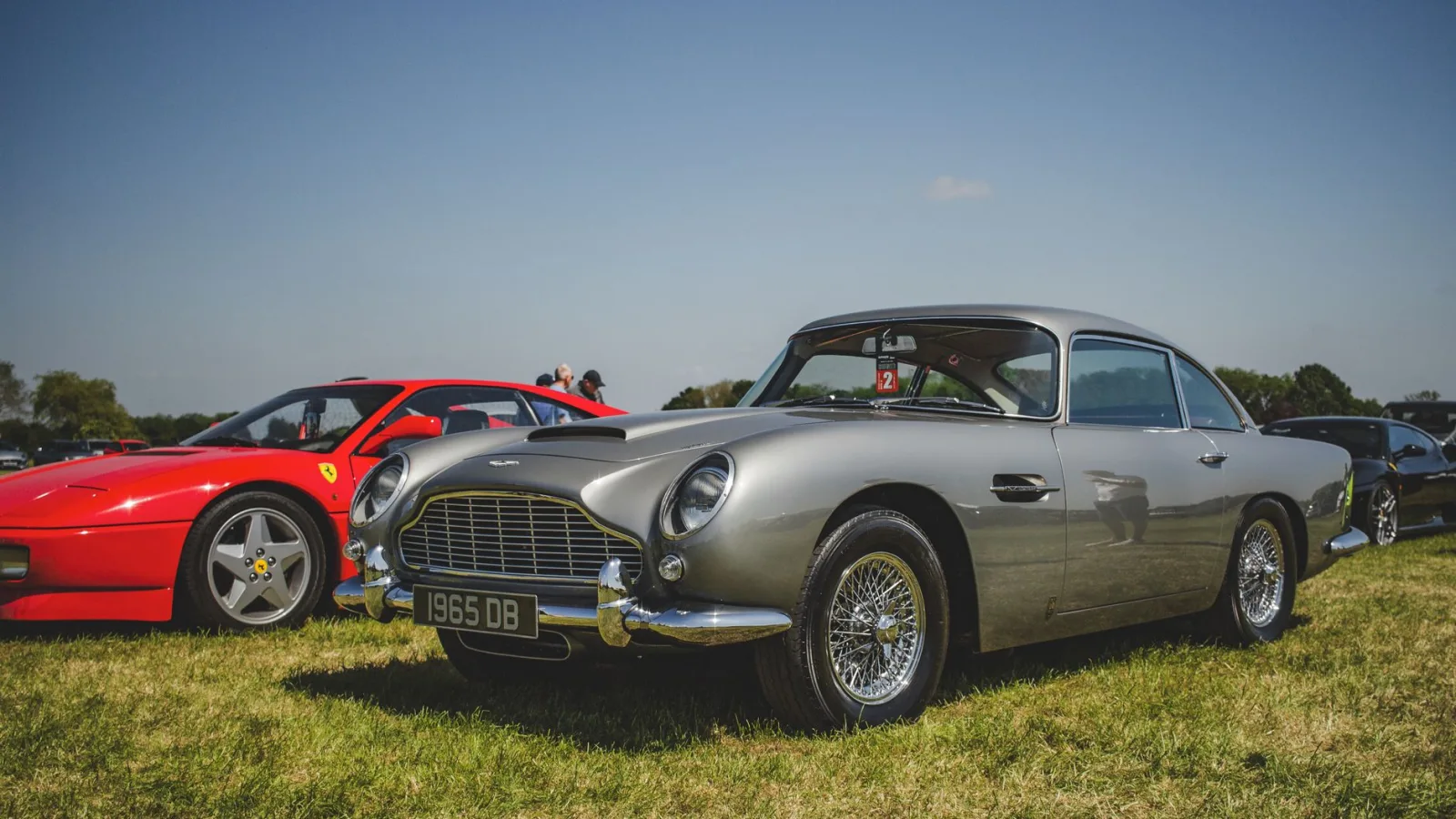 Vintage Aston Martin and red Ferrari parked on green grass under bright daylight.