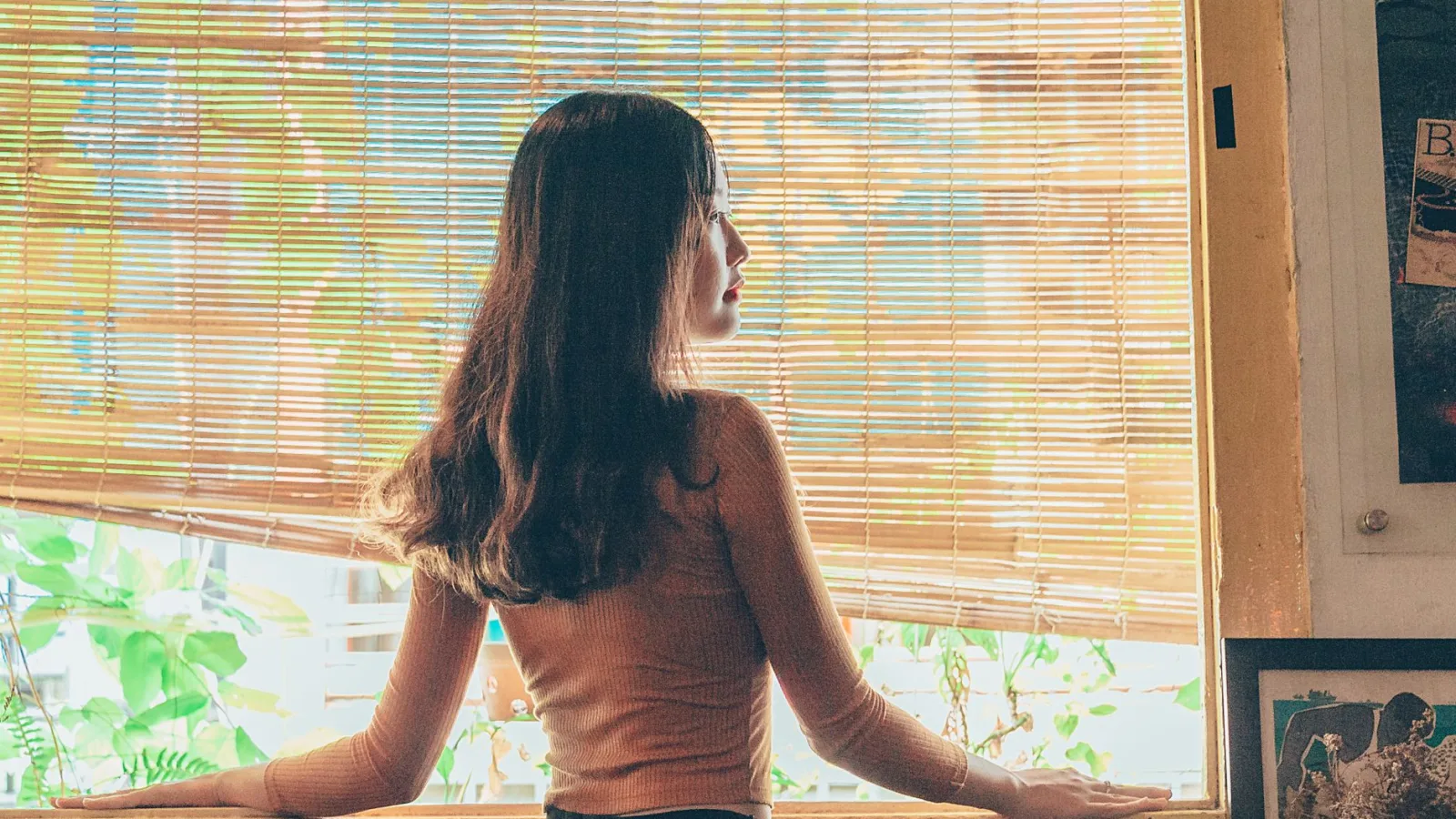 A woman stands by a window with bamboo blinds, lost in thought.