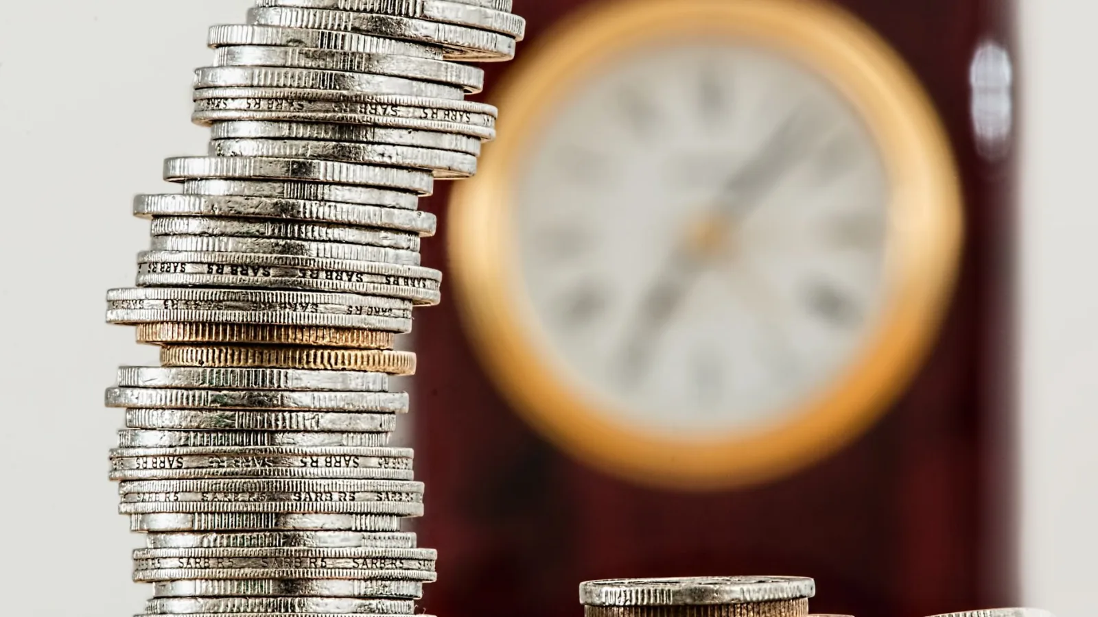 A close-up image of stacked coins with a blurred clock, symbolizing time and money relationship.