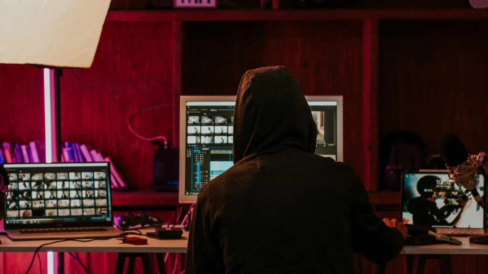 Person in a hoodie editing videos on multiple screens in a dimly lit space with neon lighting.