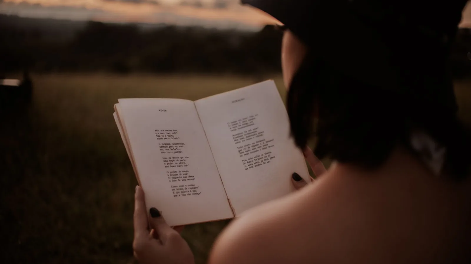 Back view of anonymous female in hat reading book with poetry in grassy field on blurred background on summer evening