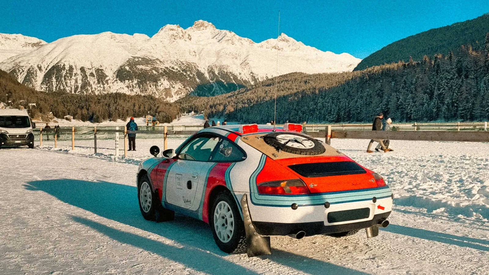 Racing car parked in a snowy mountain landscape with people enjoying winter activities.