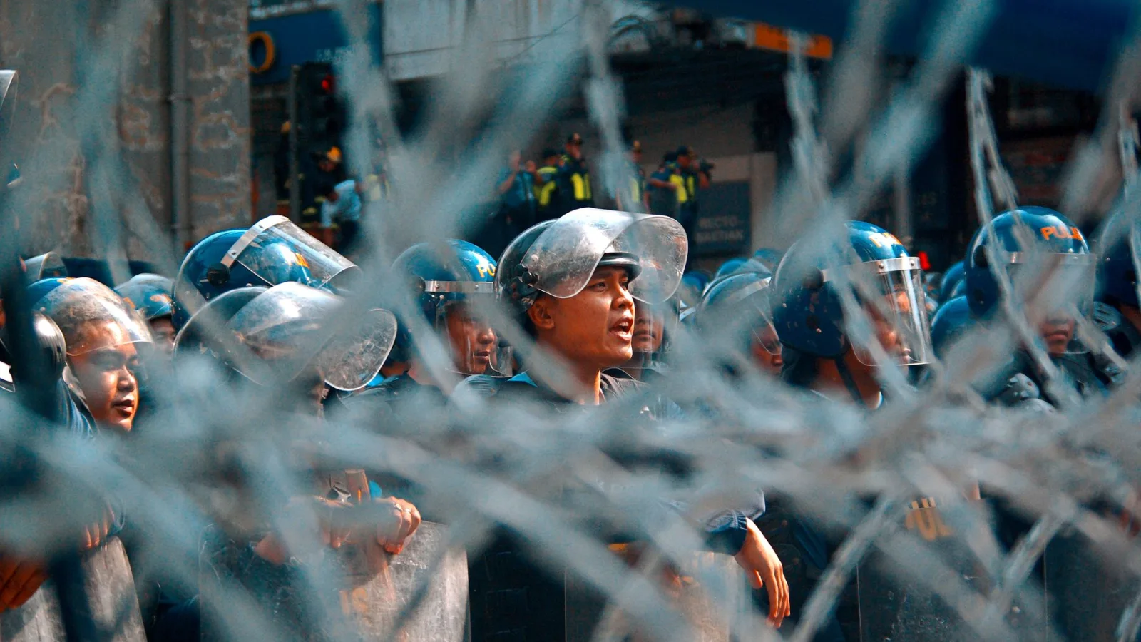 Riot police lined up behind barbed wire during a protest, showing tension and security forces.