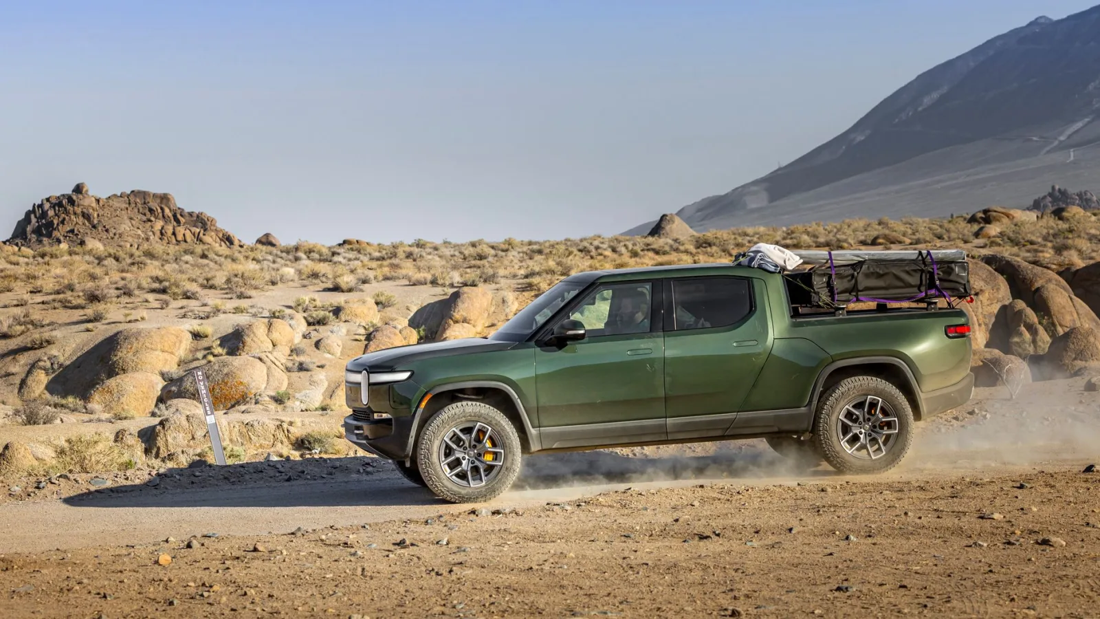 Green electric pickup truck on a dusty trail in a rocky desert landscape.