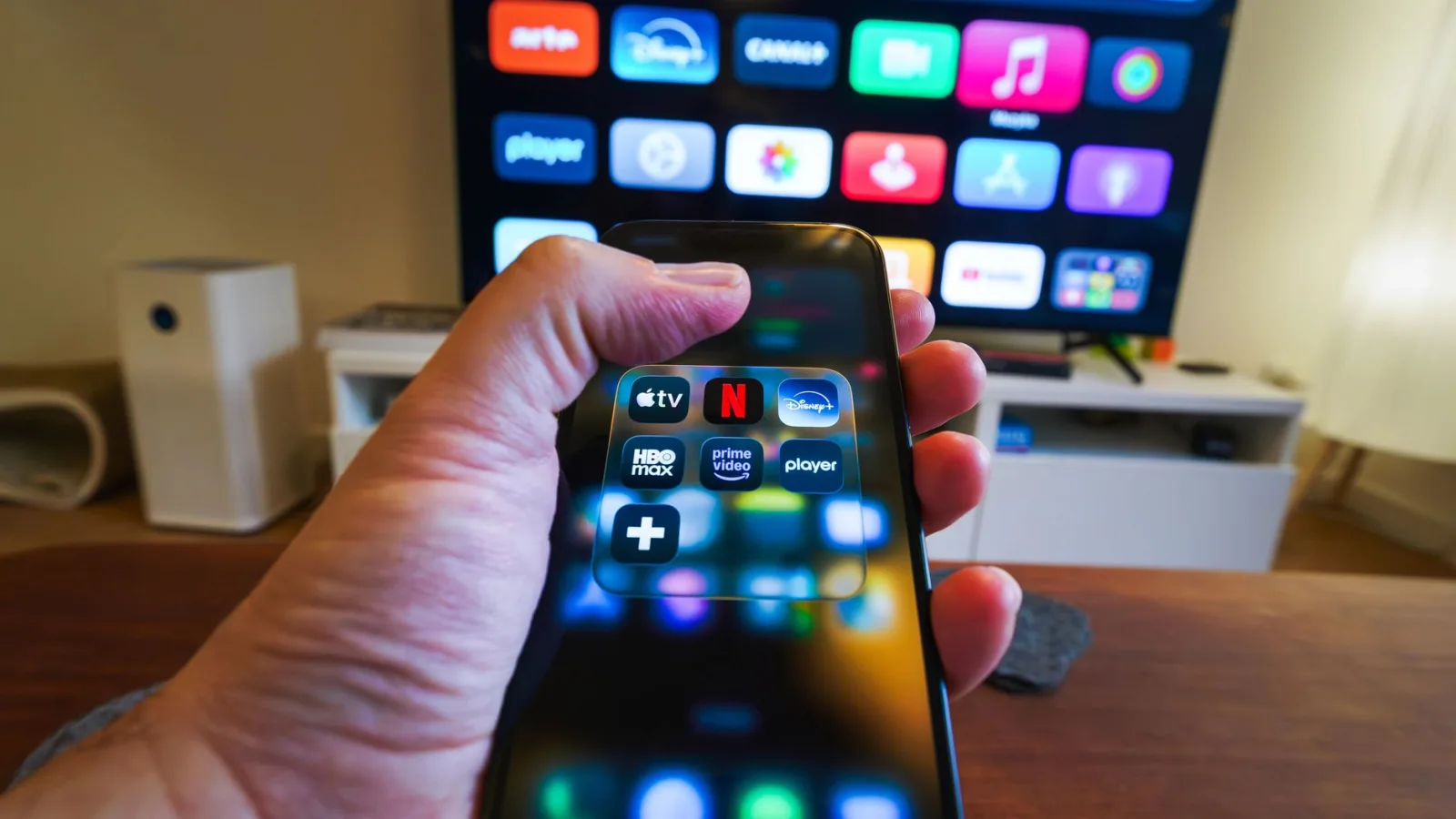 Close-up of a hand holding a phone displaying streaming apps in front of a TV with multiple app icons.