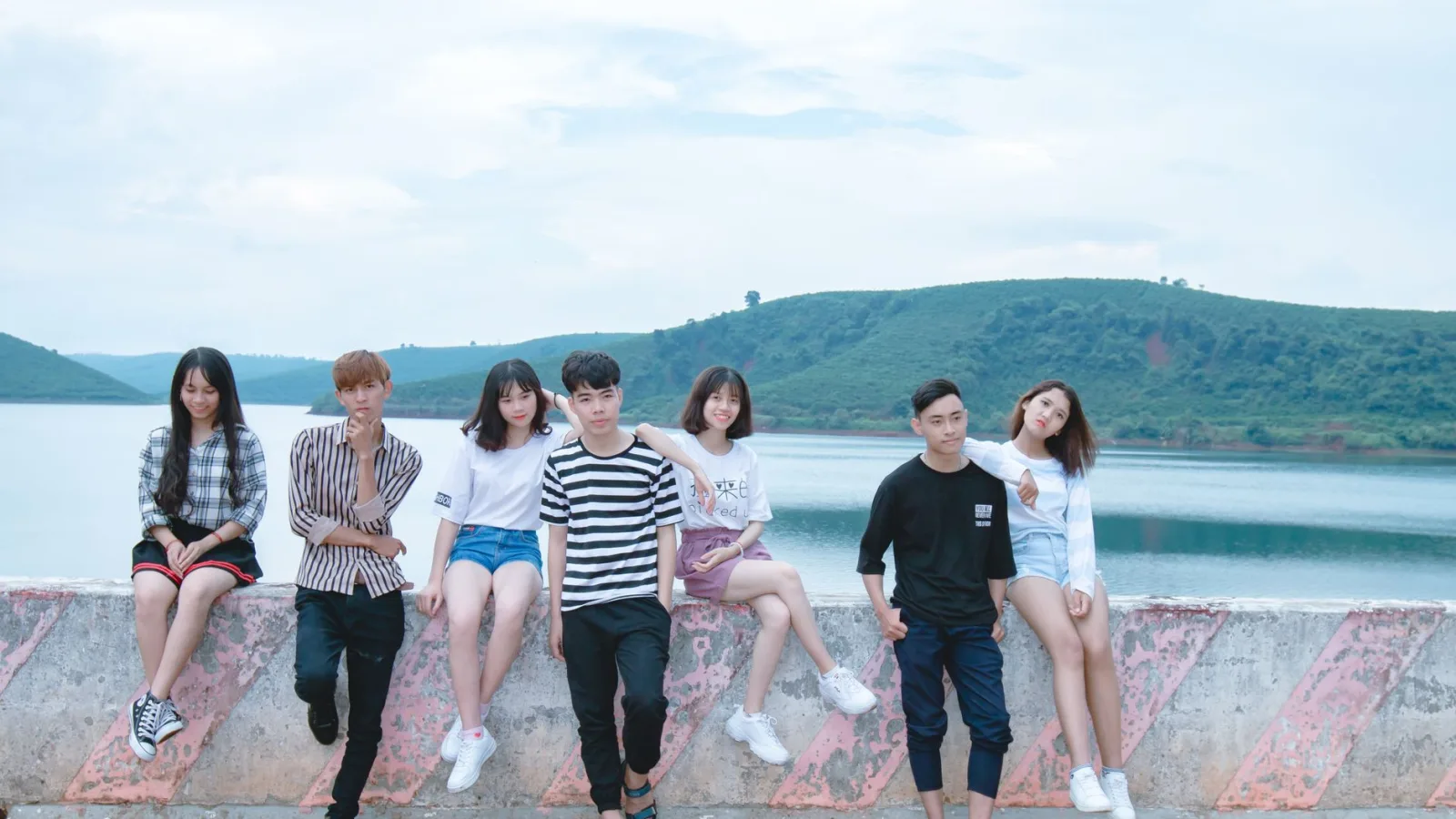 A group of young friends sitting by a serene lake with lush hills in the background, enjoying a sunny day.