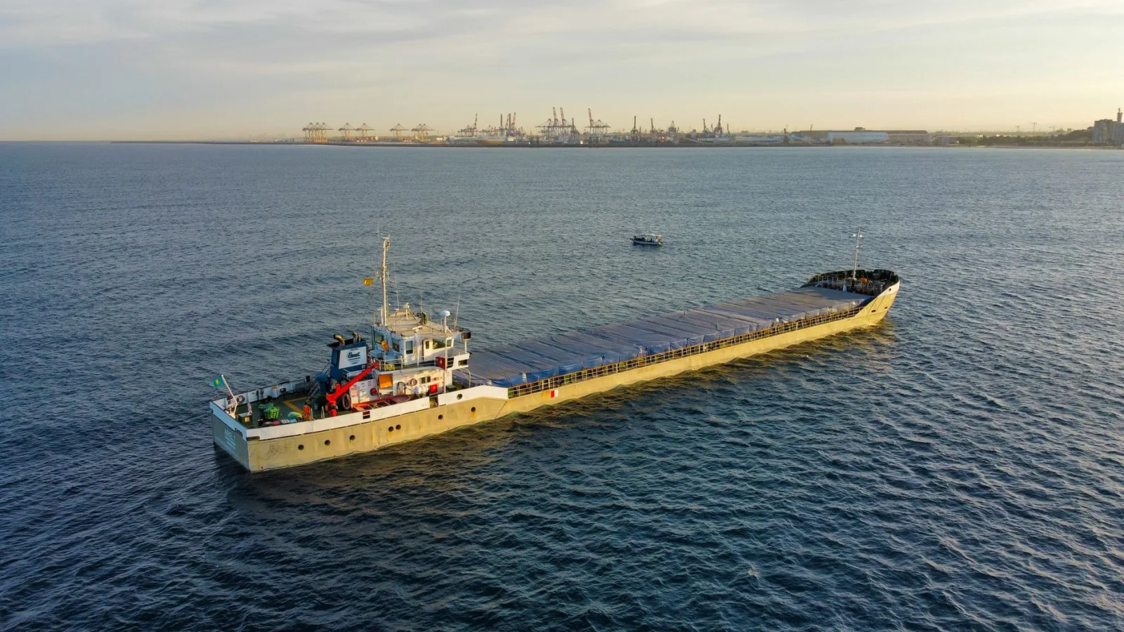 Aerial view of a cargo ship sailing near a busy industrial port at sunset.