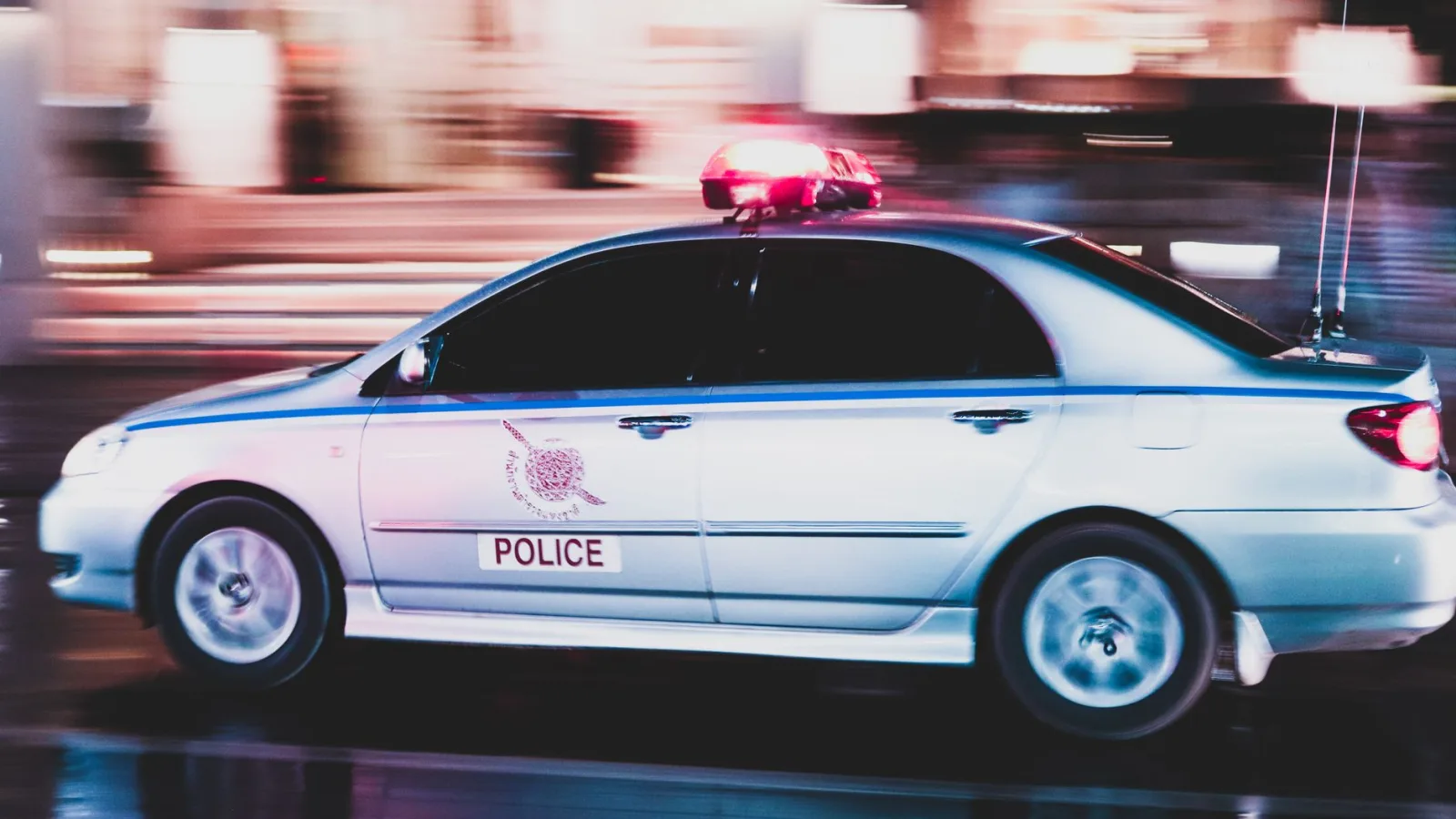A motion-blurred police car speeds through a busy street in Thailand at night.