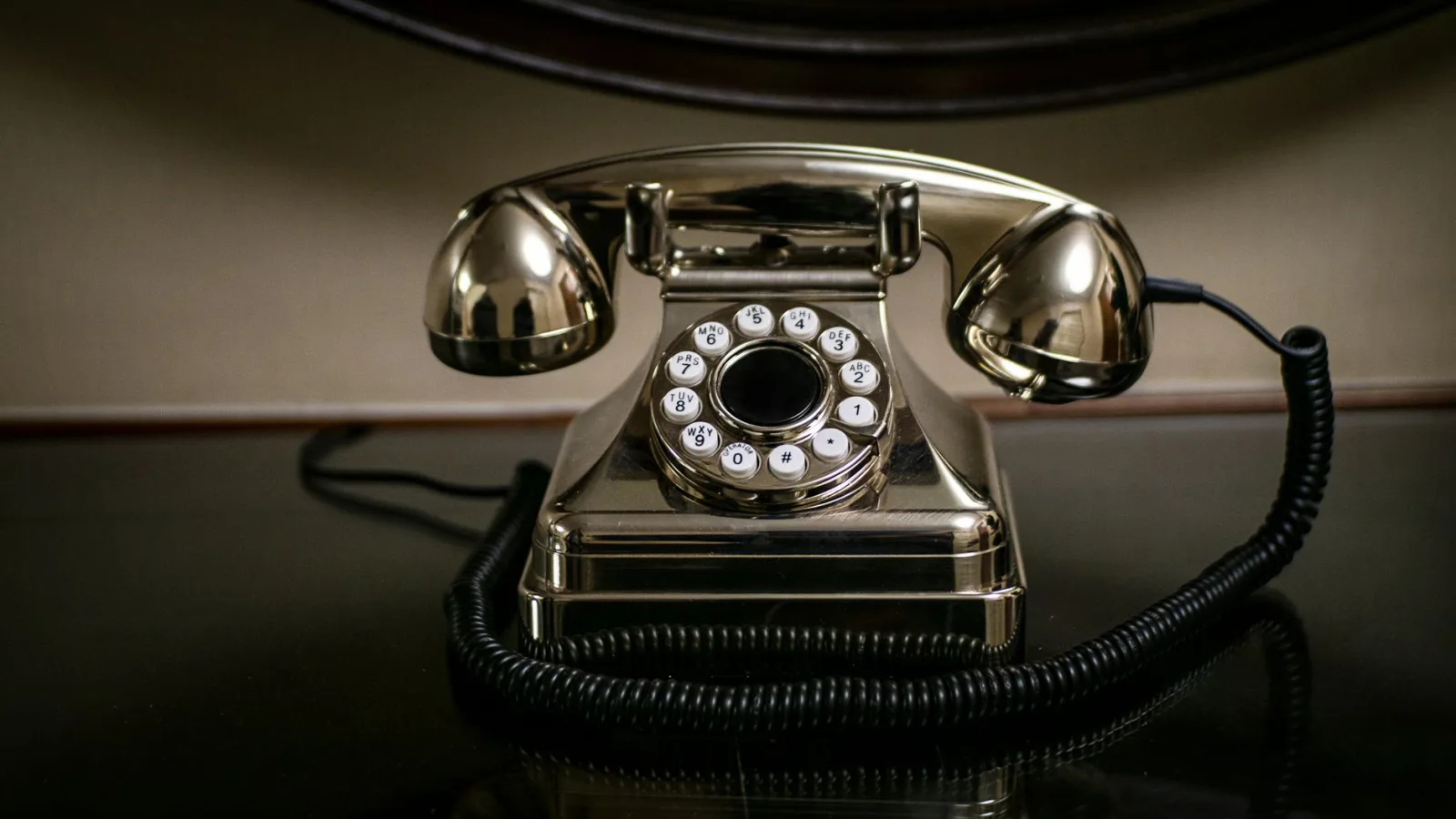 A shiny vintage dial telephone with a coiled cord on a dark reflective surface.