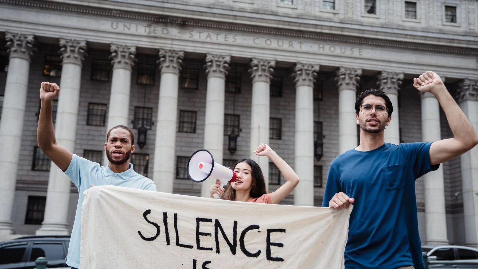 Group of diverse activists holding banner and megaphone in front of a US courthouse for a protest.